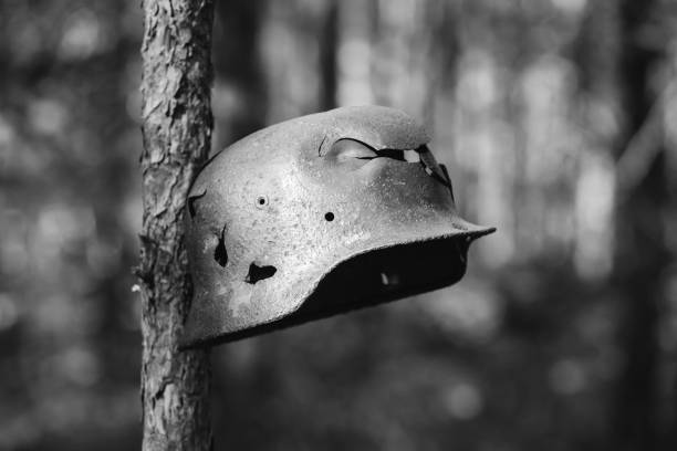 Damaged By Bullets And Shrapnel Metal Helmet Of German Infantry Wehrmacht Soldier At World War II. Rusty Helmet Hanging On Tree Trunk. Photo In Black And White Colors.